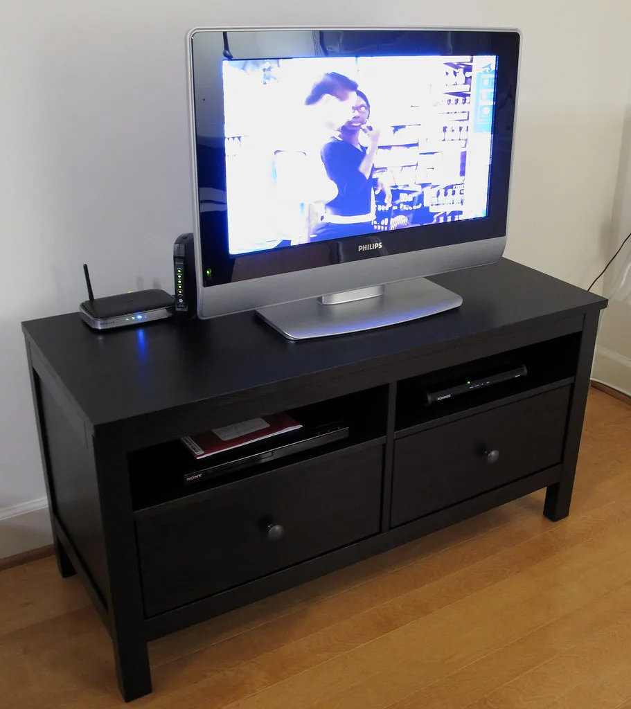 Hands measuring a TV stand shelf with a tape measure beside a flat-screen TV.