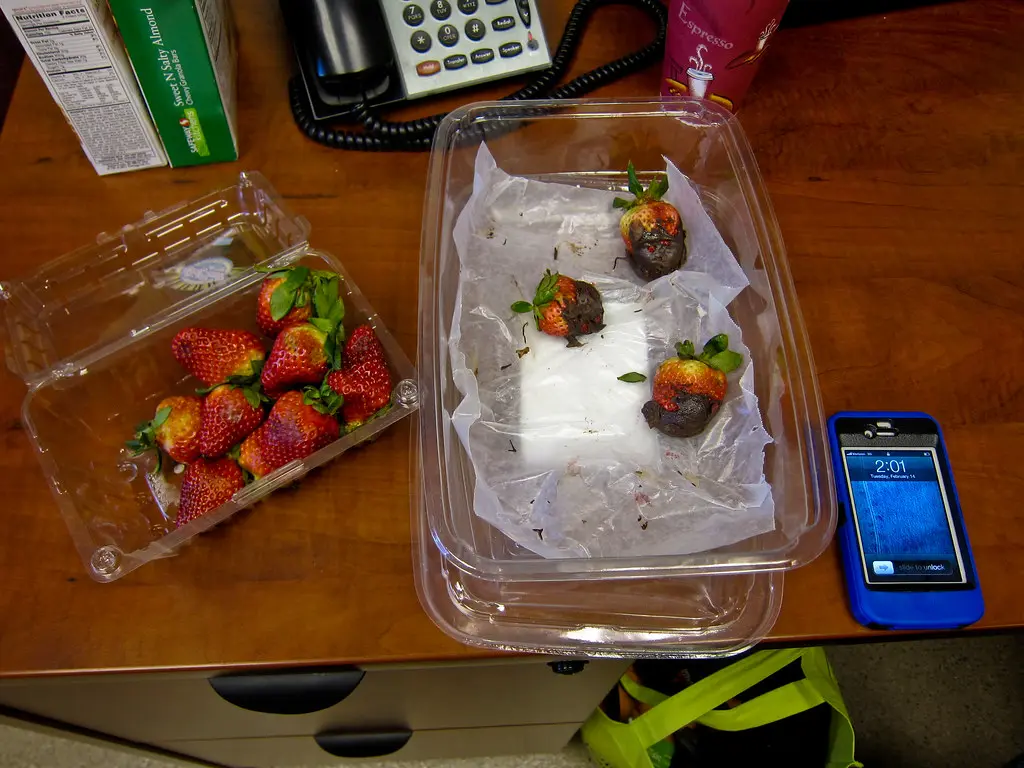 Hands dipping a strawberry into melted chocolate over a glass bowl on parchment paper