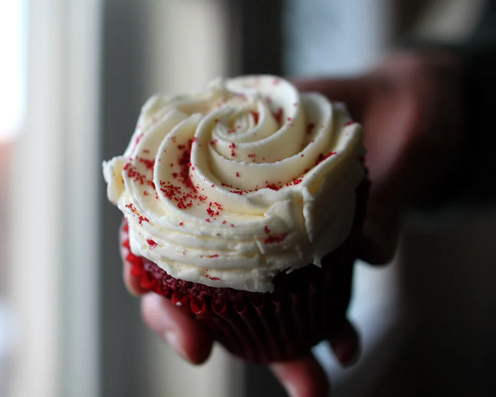 Hands piping cream cheese frosting onto red velvet cupcakes on a kitchen counter