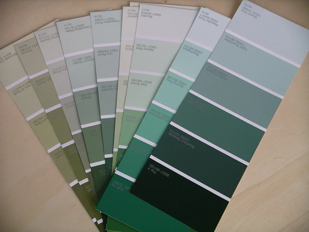 A hand holds paint swatches against a bedroom wall near a window with natural light.