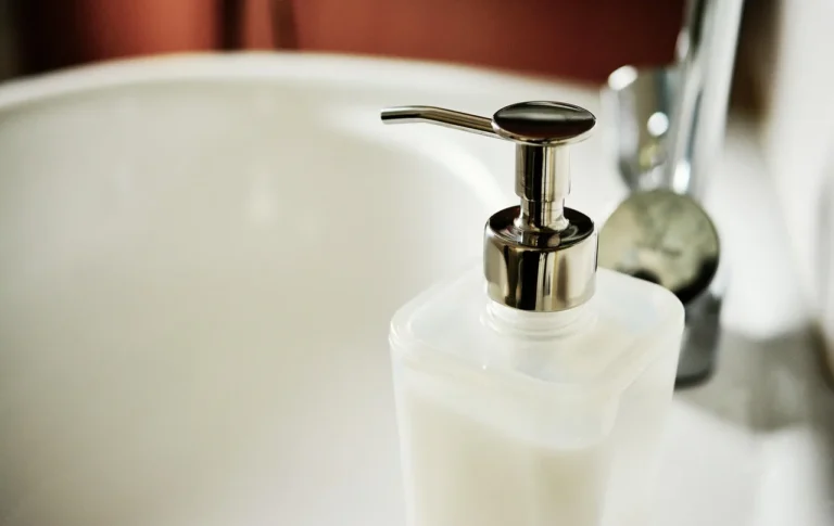 A person wipes a bathroom sink and countertop in natural light with a spray bottle nearby.